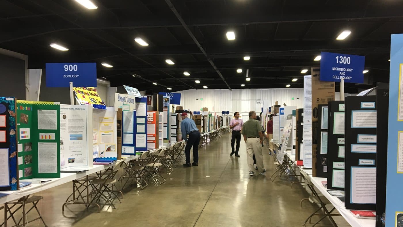An aisle of the exhibit hall at the Orange County Science and Engineering Fair, project boards on both sides under category signs for zoology and microbiology.