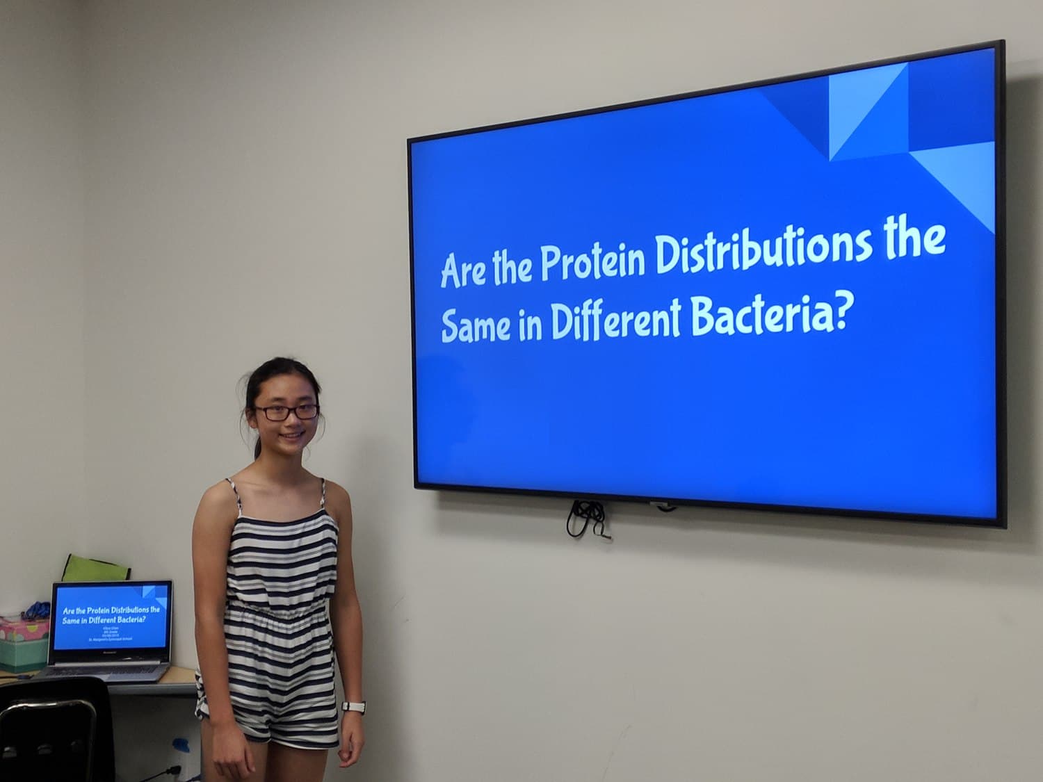 An Ardent Scholar presenting her experimental design beside a slide reading “Are the protein distributions the same in different bacteria?”